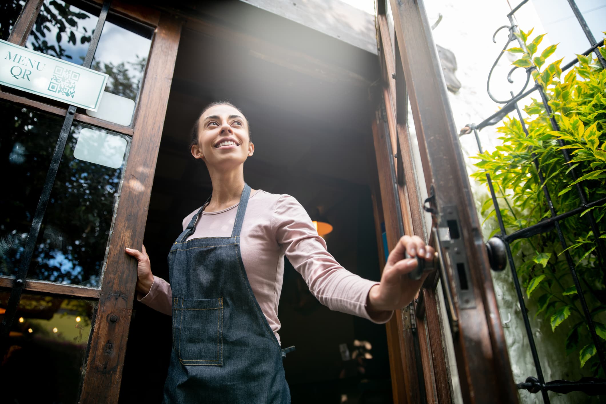 Una persona sonriente, con un delantal, está de pie junto a la puerta abierta de una cafetería o un restaurante, mirando hacia fuera. La luz del sol entra por la puerta y junto a ella se ve un menú con un código QR. A la derecha se ven plantas verdes.