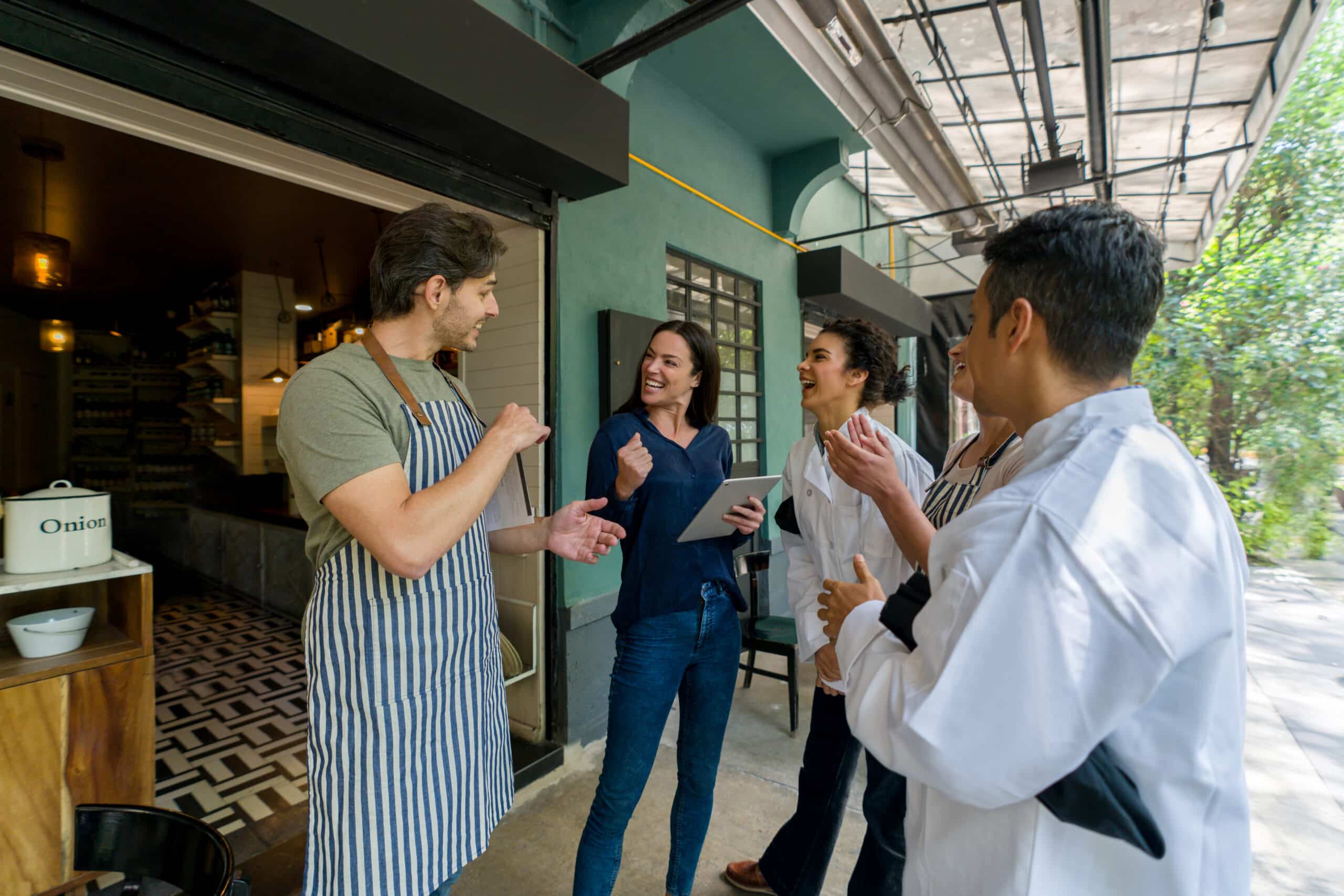 Cuatro personas, dos de ellas con delantal, están de pie charlando animadamente a la puerta de un restaurante. Una de ellas sostiene una tableta, y todos parecen estar disfrutando de la animada conversación en un día soleado.