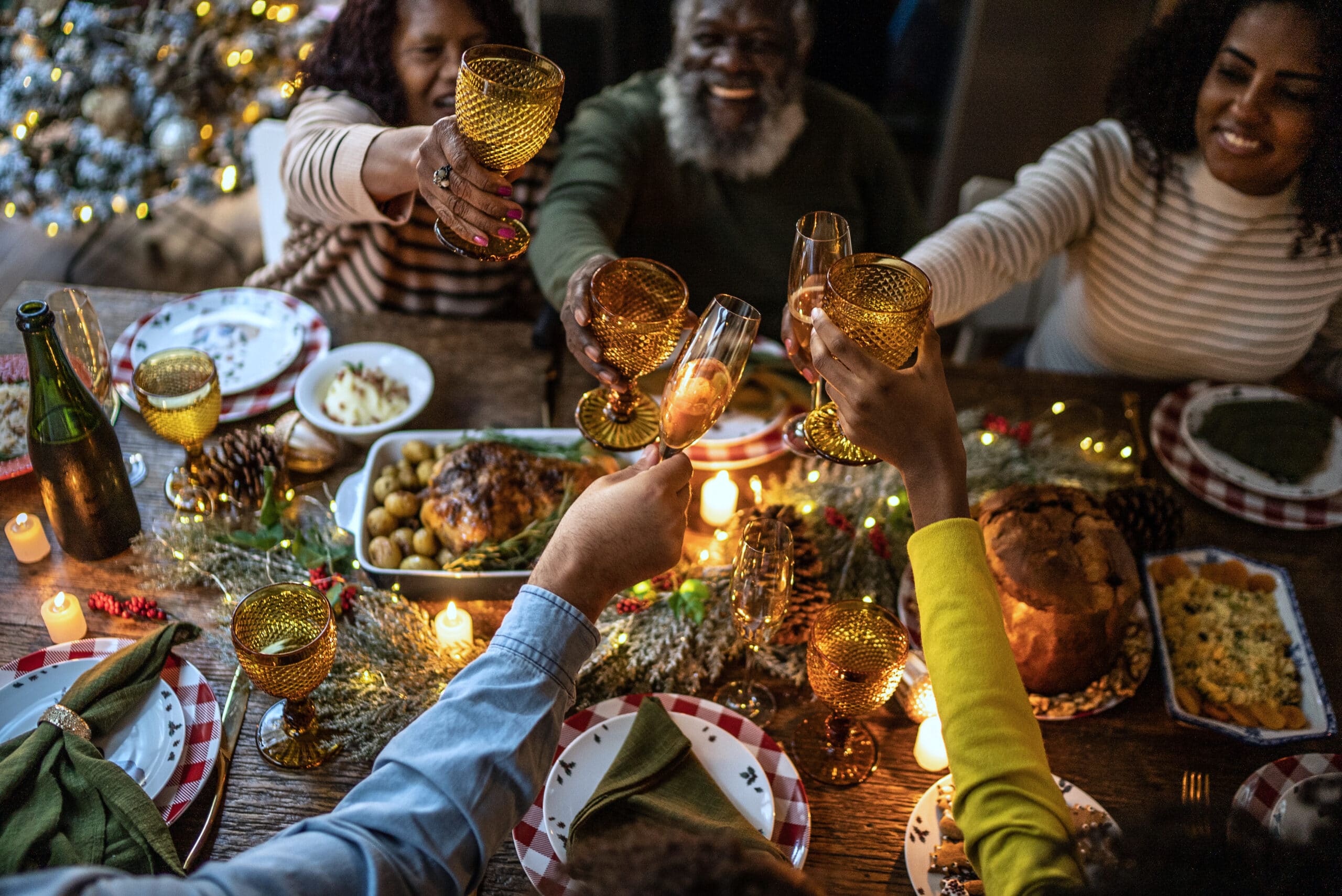 Un grupo de personas está sentado alrededor de una mesa festiva, levantando las copas para brindar. La mesa está decorada con velas, adornos navideños de hojas verdes y diversos platos. Al fondo se ve un árbol de Navidad difuminado.