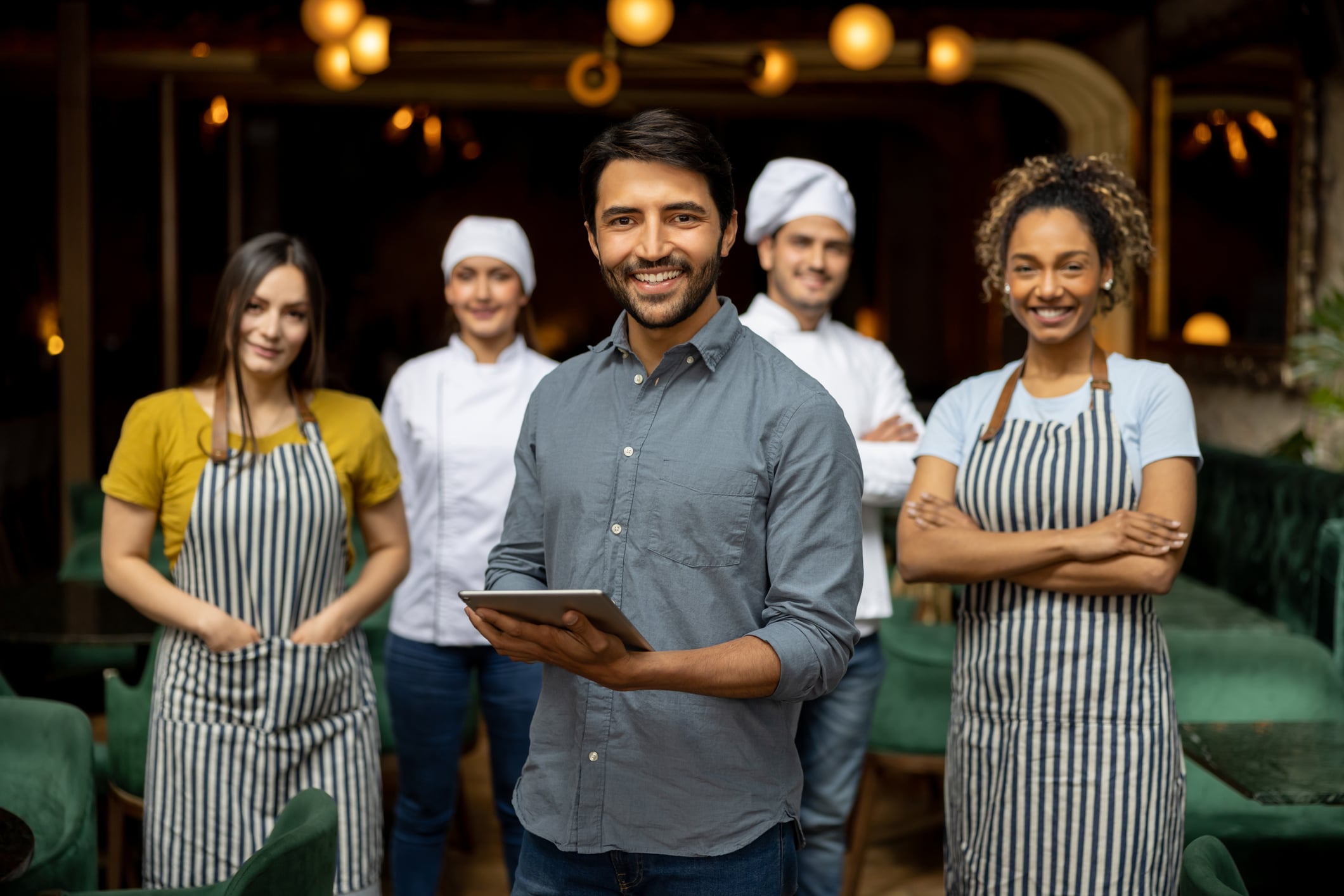 Un hombre sonriente, con una tableta en la mano, está de pie frente a cuatro empleados del restaurante que llevan delantales y gorros de cocinero, posando juntos en el interior de un restaurante con una iluminación cálida.