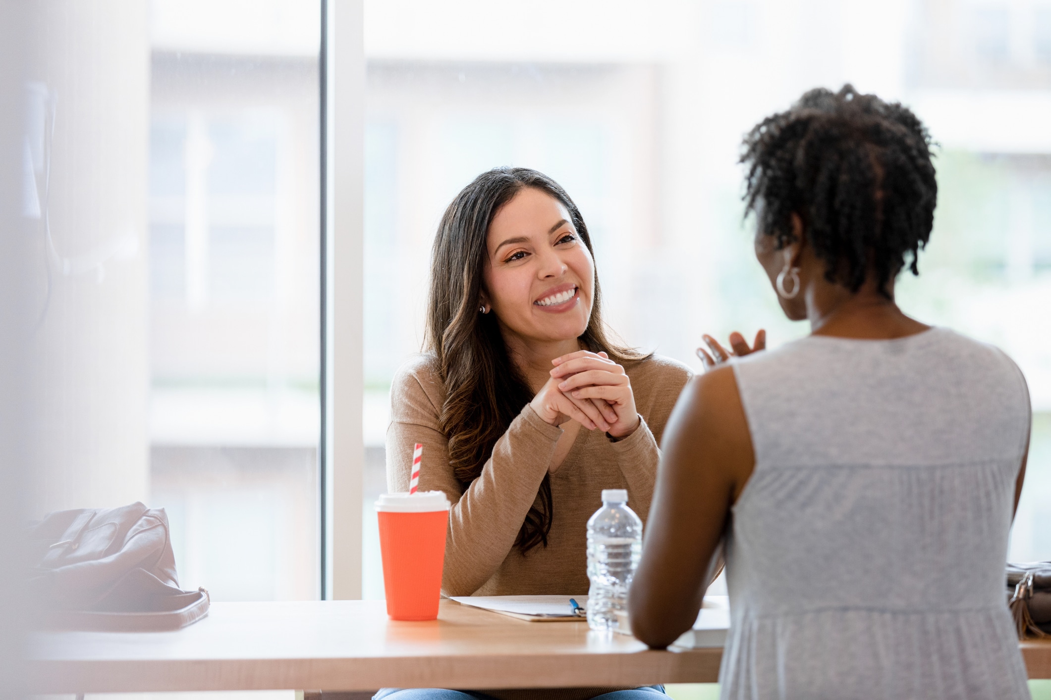 Dos mujeres están sentadas en una mesa junto a una ventana, sonriendo y charlando. Una tiene un cuaderno y un vaso rojo con una pajita; la otra sostiene una botella de agua. El ambiente parece distendido y acogedor.