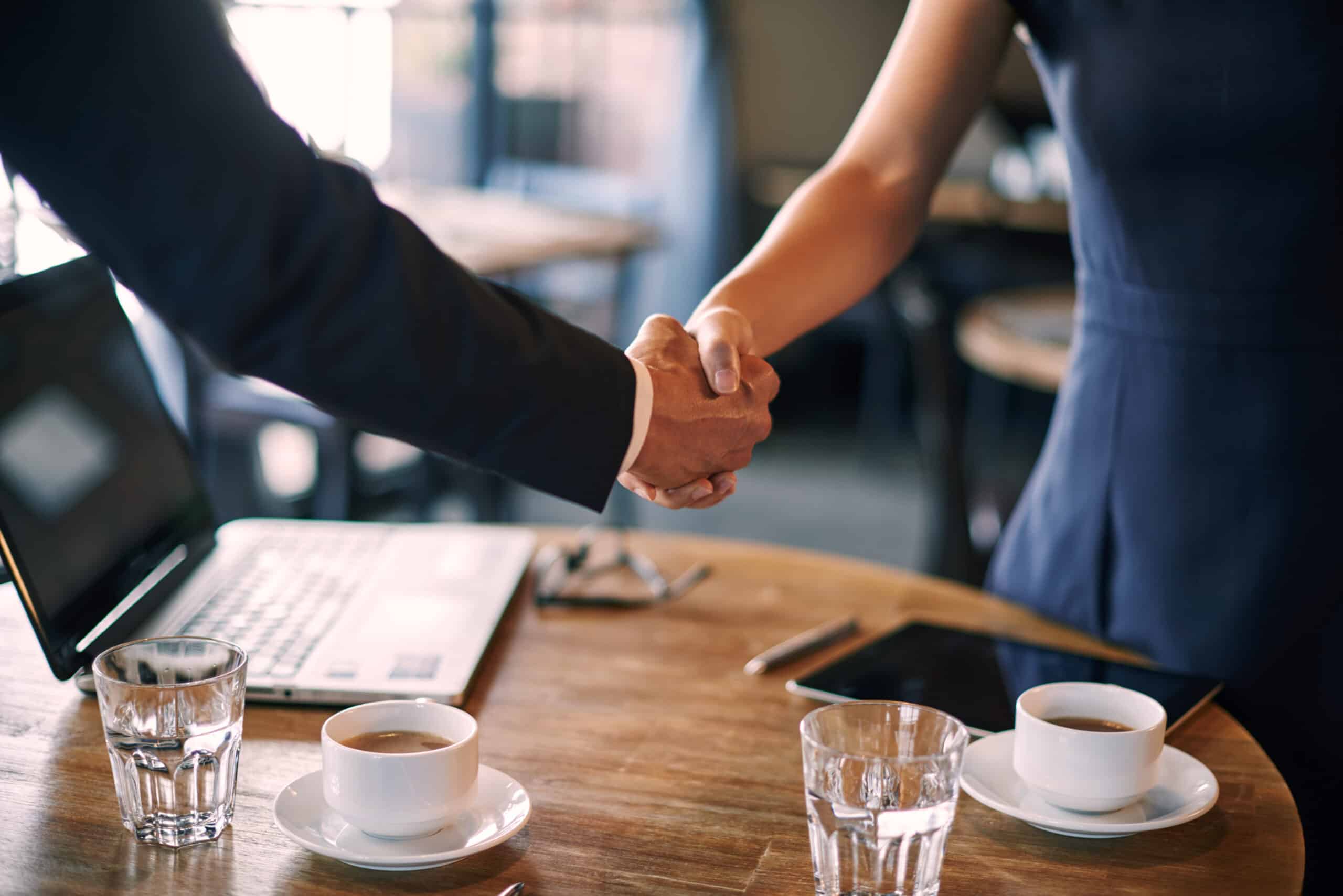 Dos personas se dan la mano al otro lado de una mesa de madera en la que hay tazas de café, vasos de agua, un ordenador portátil y una tableta, lo que sugiere una reunión de negocios o un acuerdo en un ambiente informal.