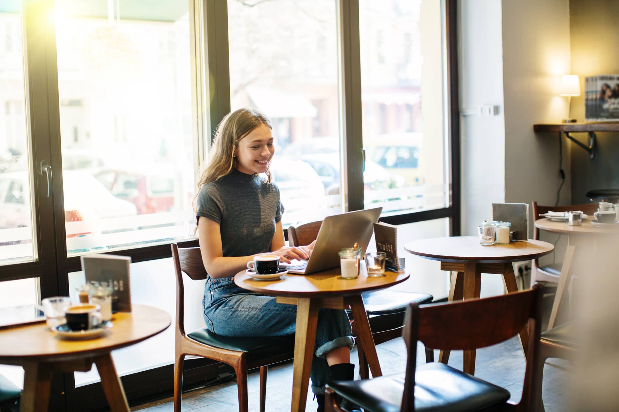 Una joven está sentada en una mesa de una acogedora cafetería, sonriendo mientras trabaja con su ordenador portátil. La luz del sol entra a raudales por los grandes ventanales y hay tazas de café sobre las mesas que la rodean.