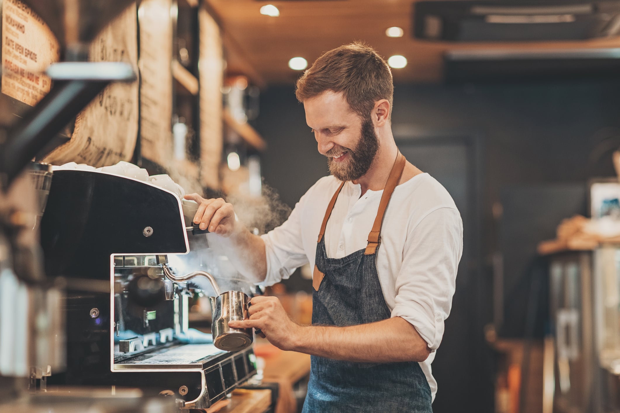 Un barista con barba y delantal sonríe mientras monta espuma de leche con la boquilla de vapor de una cafetera en una acogedora cafetería.