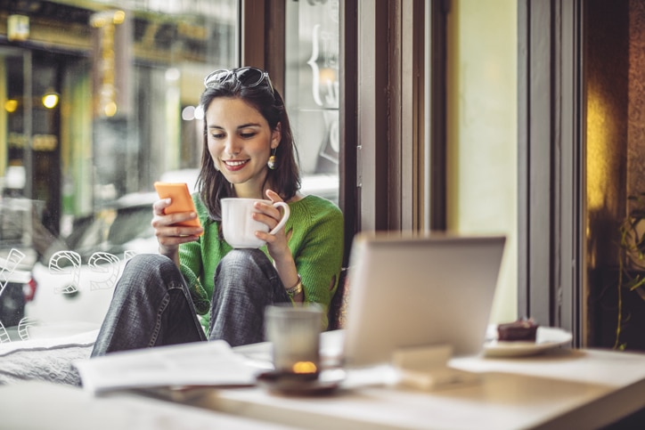 Una mujer con un jersey verde está sentada junto a la ventana de una cafetería, sonriendo a su móvil mientras sostiene una taza. Sobre la mesa que tiene delante hay un ordenador portátil abierto, un cuaderno, una vela y un postre.