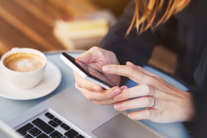Primer plano de las manos de una persona que utiliza un smartphone junto a un portátil abierto y una taza de café sobre una mesa.