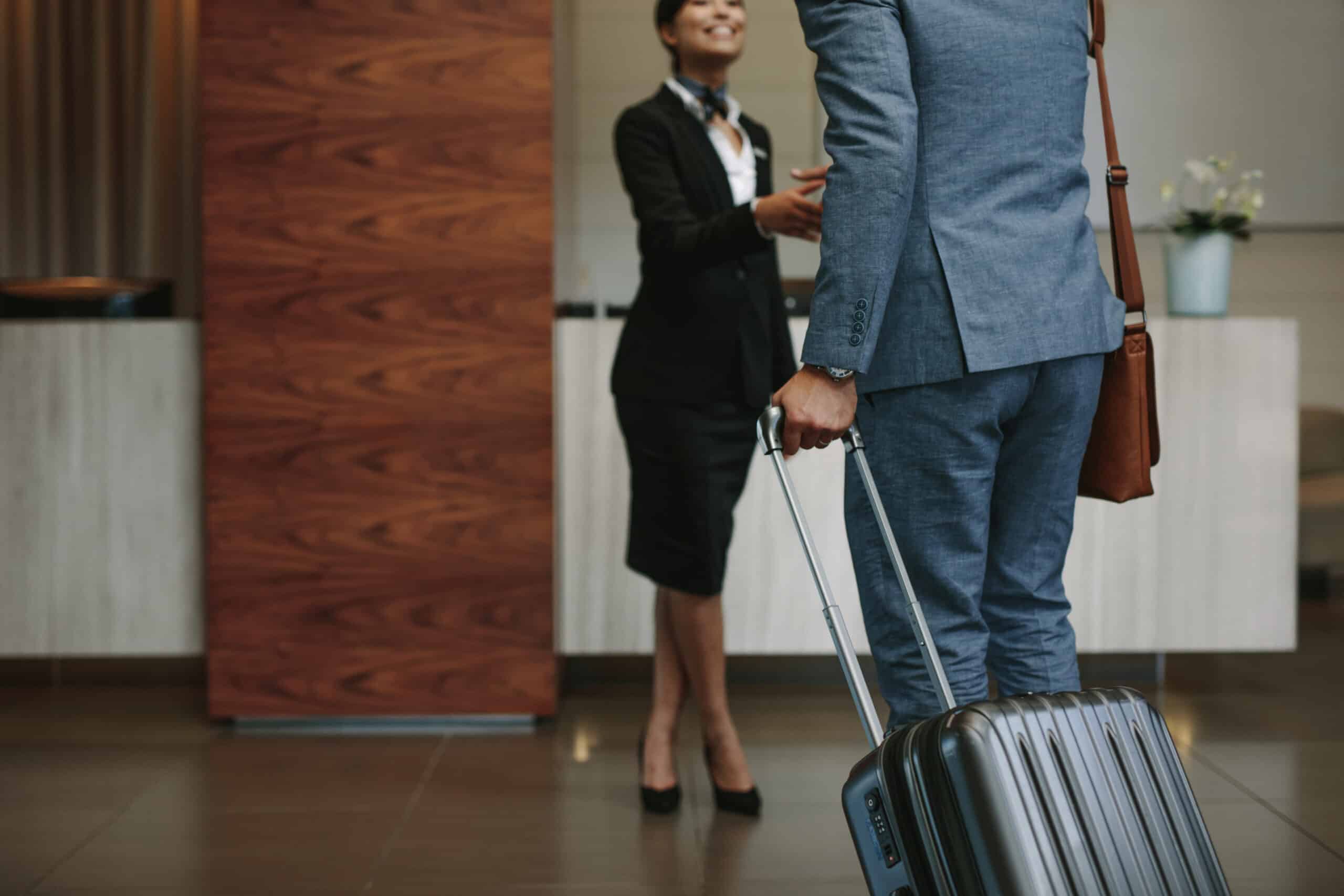 Una persona vestida con traje y con una maleta con ruedas en la mano está de pie en el mostrador de recepción de un hotel, mientras que una recepcionista, vestida con ropa de oficina, le sonríe y le da la mano.