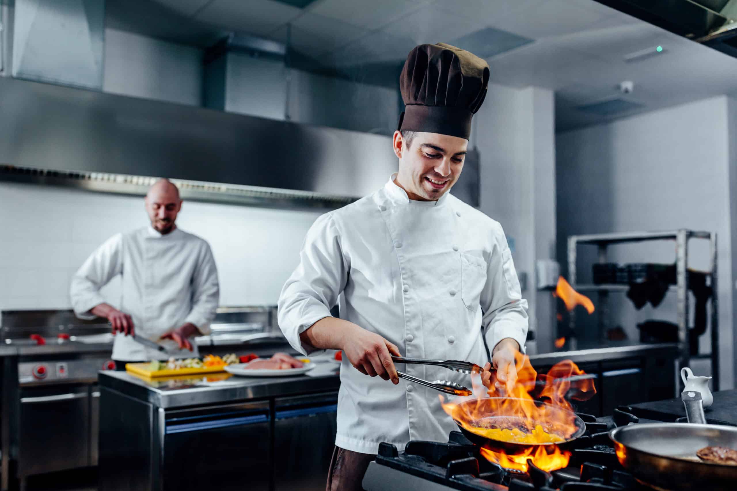 Un cocinero, vestido con un uniforme blanco y un gorro, cocina con las llamas de una sartén sobre una cocina, sonriendo, mientras otro cocinero trabaja al fondo en una cocina profesional.