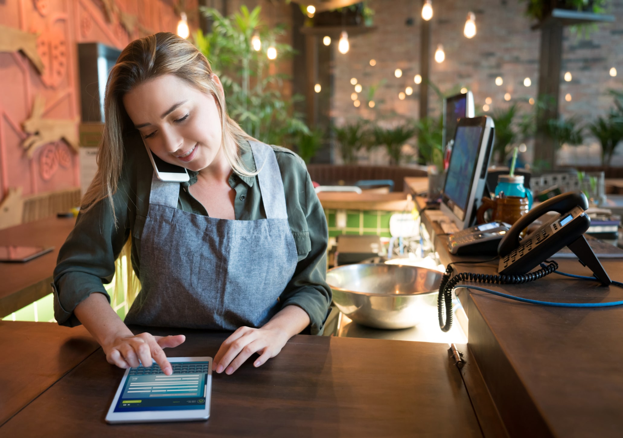 Una mujer con delantal realiza varias tareas a la vez en una barra, con el teléfono pegado a la oreja mientras utiliza una tableta. Parece estar trabajando en una cafetería o un restaurante moderno, con plantas e iluminación cálida al fondo.