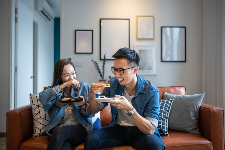 Dos personas están sentadas en un sofá marrón, sonriendo y comiendo pizza juntas en un acogedor salón con cuadros enmarcados en la pared detrás de ellas.