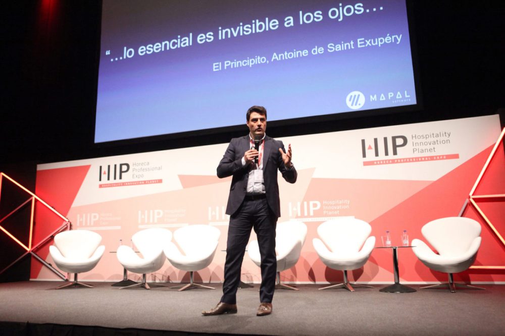 Un hombre vestido con traje habla en el escenario de una conferencia, con sillas vacías a su lado. En una gran pantalla se muestra una cita en español de «El Principito» y, al fondo, se ve el logotipo de la conferencia.