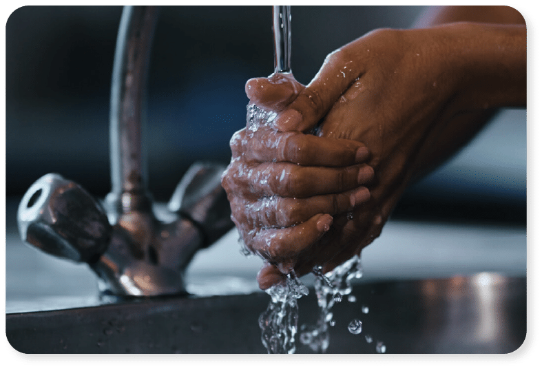 Primer plano de unas manos que se lavan bajo el chorro de agua corriente de un grifo de cocina, con el agua salpicando sobre las manos y cayendo en un fregadero metálico.