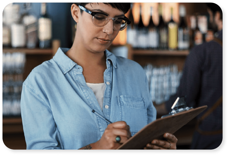 Una persona que lleva gafas y una camisa vaquera escribe en un portapapeles en un bar o restaurante, con botellas y vasos visibles al fondo.