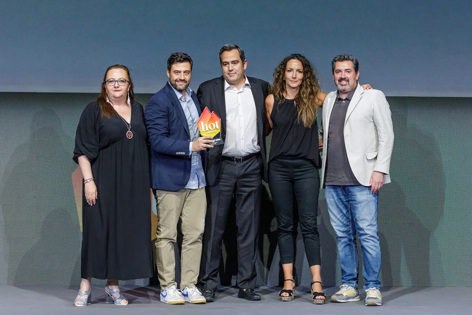 Cinco personas están de pie juntas en un escenario; algunas sonríen, y una de ellas, en el centro, sostiene un premio rojo y amarillo en el que se lee «hot». Visten ropa de negocios o informal elegante. El fondo es gris liso.