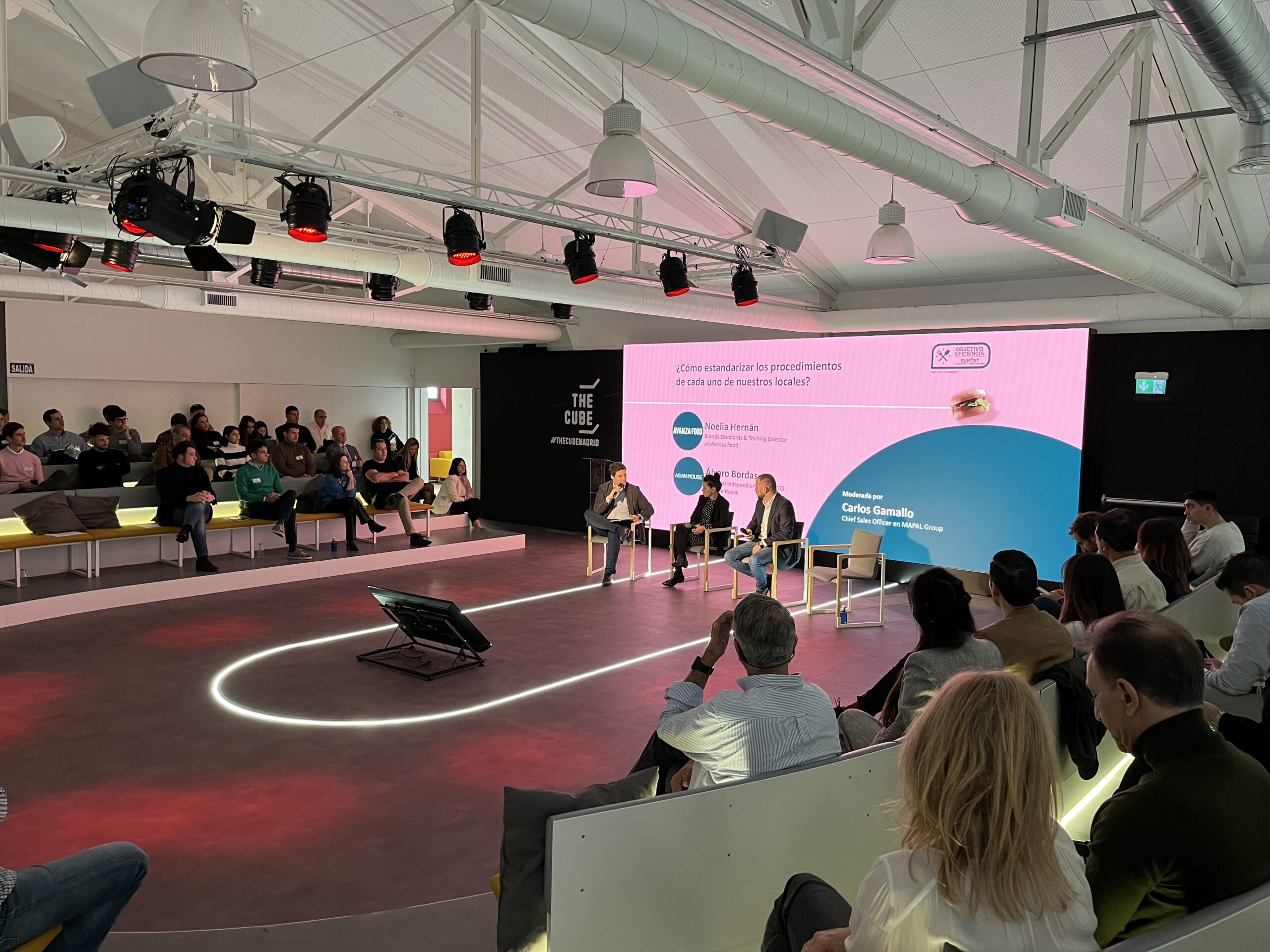 En un escenario tiene lugar una mesa redonda en la que dos ponentes están sentados bajo los focos, mientras el público escucha. En una gran pantalla se muestra el contenido de la presentación en español. El escenario es moderno, con iluminación de estilo industrial.