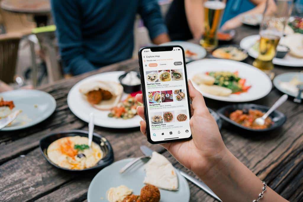 Una persona sostiene un smartphone en el que se ve una aplicación de reparto de comida, mientras está sentada en una mesa de madera con platos de diversos manjares y bebidas, rodeada de gente que comparte una comida.