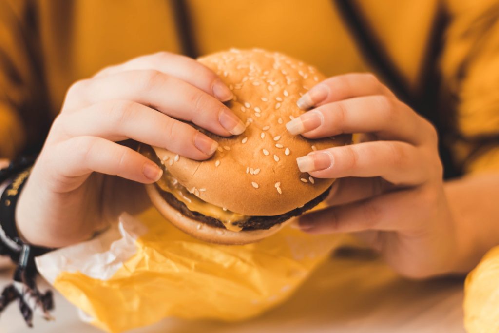 Una persona que sostiene con ambas manos una hamburguesa con semillas de sésamo y la está desenvolviendo del papel amarillo.