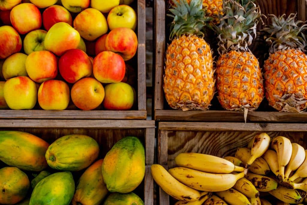 Cajas de madera llenas de manzanas, piñas, papayas y plátanos dispuestas en un puesto del mercado. Las frutas están ordenadas por tipo, lo que crea una presentación colorida y ordenada.