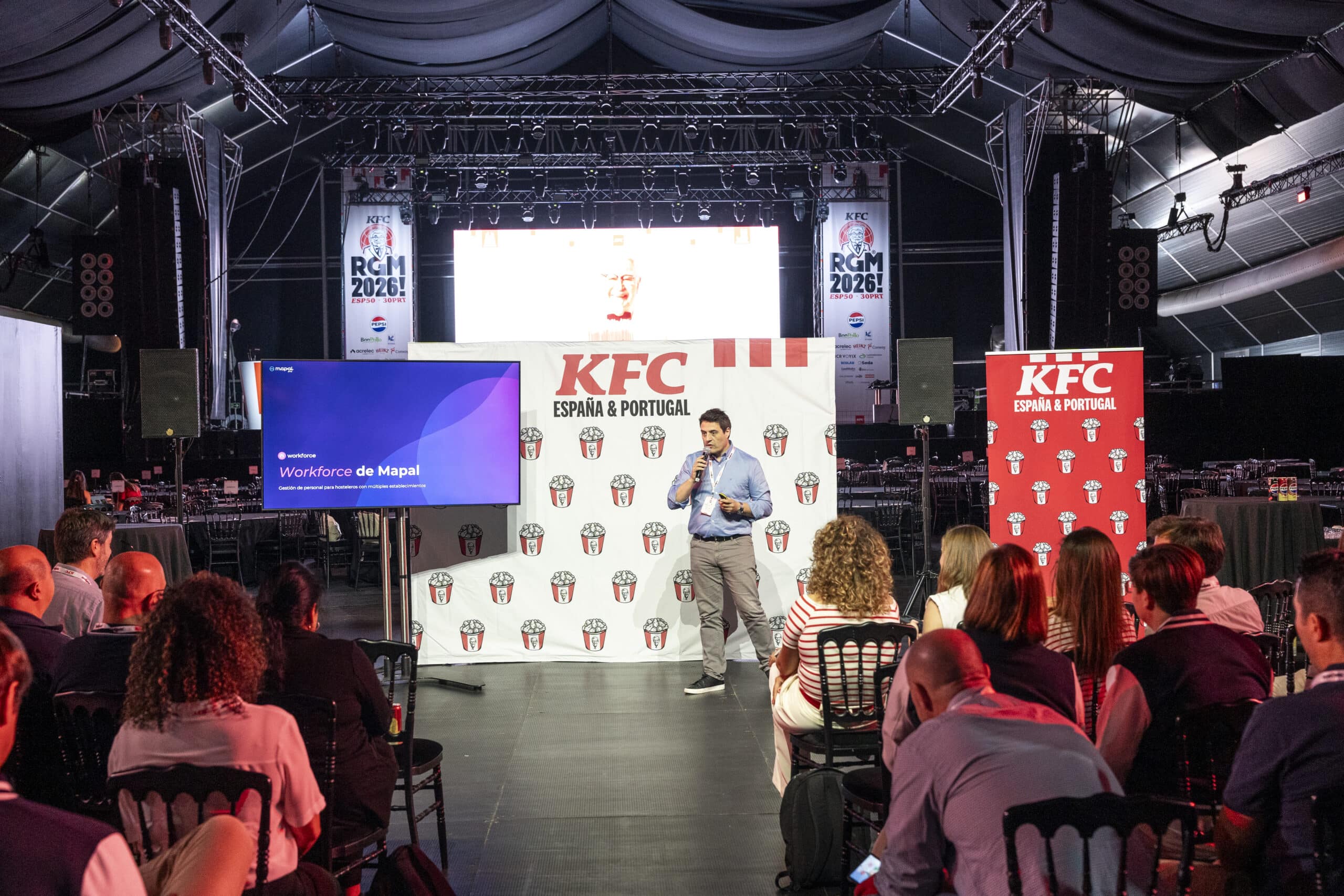 Un hombre se encuentra de pie en el escenario dando una presentación ante el público en un evento de KFC España y Portugal, con una pantalla en la que se ven «Workforce de Mapal» y carteles de KFC al fondo, en el interior de un recinto amplio y con poca luz.