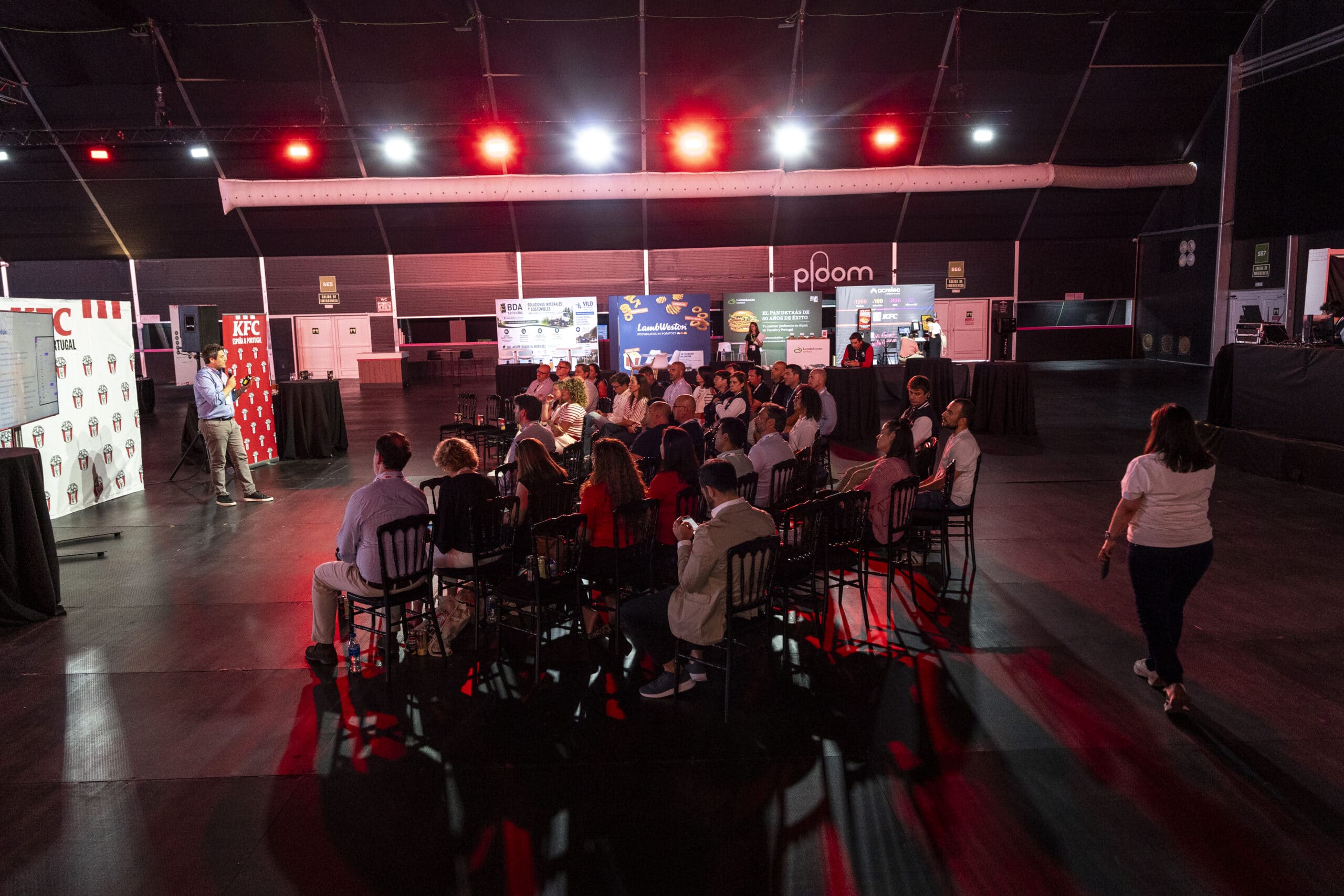 Un grupo de personas está sentado en filas, de cara a un orador que se encuentra en un escenario, bajo luces rojas en un gran recinto cubierto con carteles publicitarios de la marca al fondo.