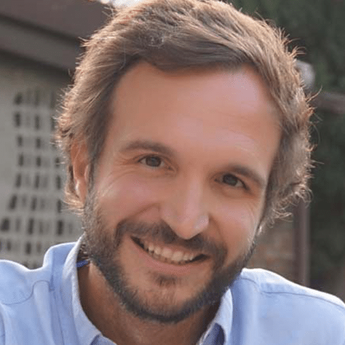 Un hombre con el pelo castaño claro y barba, vestido con una camisa azul claro, sonriendo al aire libre con vegetación y una estructura borrosa al fondo.