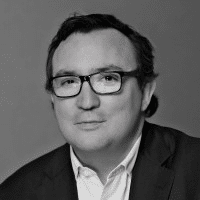 Un hombre de pelo oscuro, con gafas, chaqueta de traje y camisa blanca con cuello, posa ante un fondo liso. La imagen es en blanco y negro.