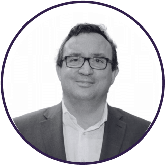 Un hombre de pelo corto y oscuro y con gafas, vestido con chaqueta de traje y camisa de cuello, sonríe a la cámara. La imagen es en blanco y negro y está enmarcada en un borde circular.