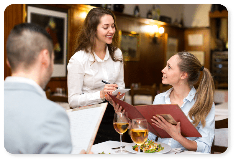 Una camarera toma nota del pedido de una mujer que sostiene una carta de menú en una mesa de restaurante, mientras otra persona está sentada cerca. Sobre la mesa hay dos copas de vino blanco y unos platos con comida.