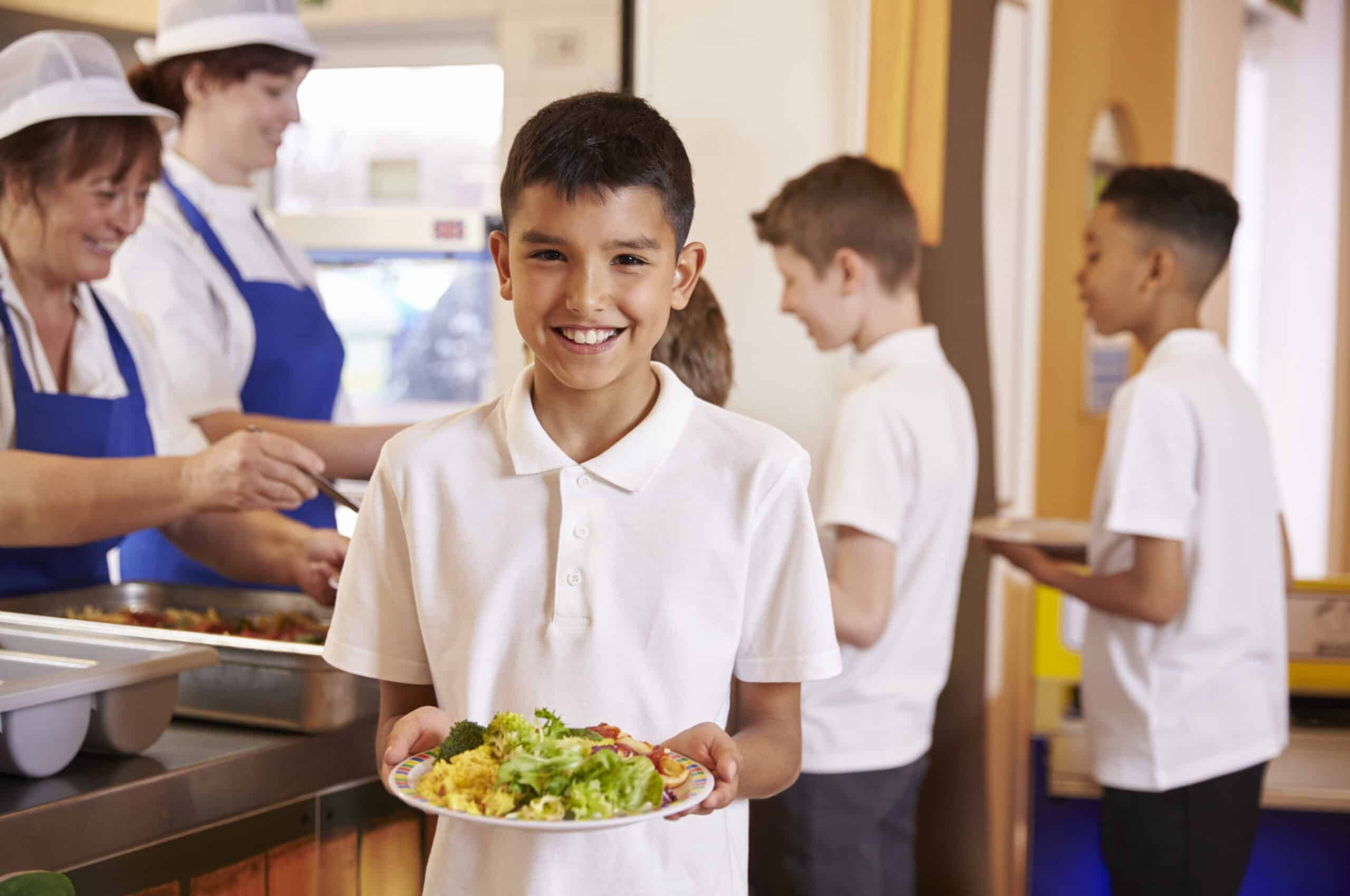 Un niño con un polo blanco sonríe mientras sostiene un plato de comida en la cola del comedor; detrás de él, otros niños esperan a que los empleados del comedor les sirvan la comida.