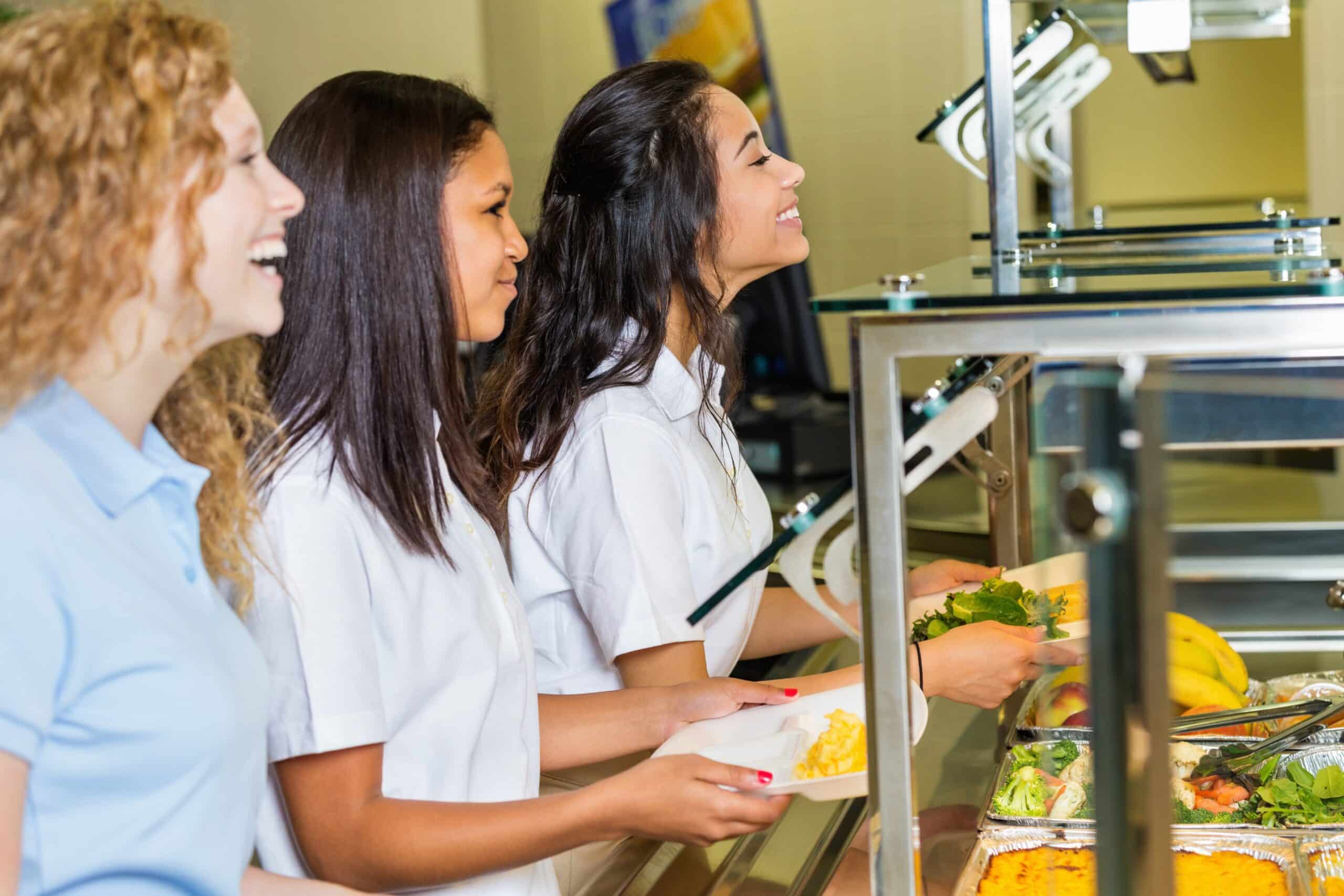 Tres chicas jóvenes hacen cola en una cafetería, sonriendo mientras eligen platos del mostrador; cada una sostiene una bandeja en la que se ven diversas opciones de comida.