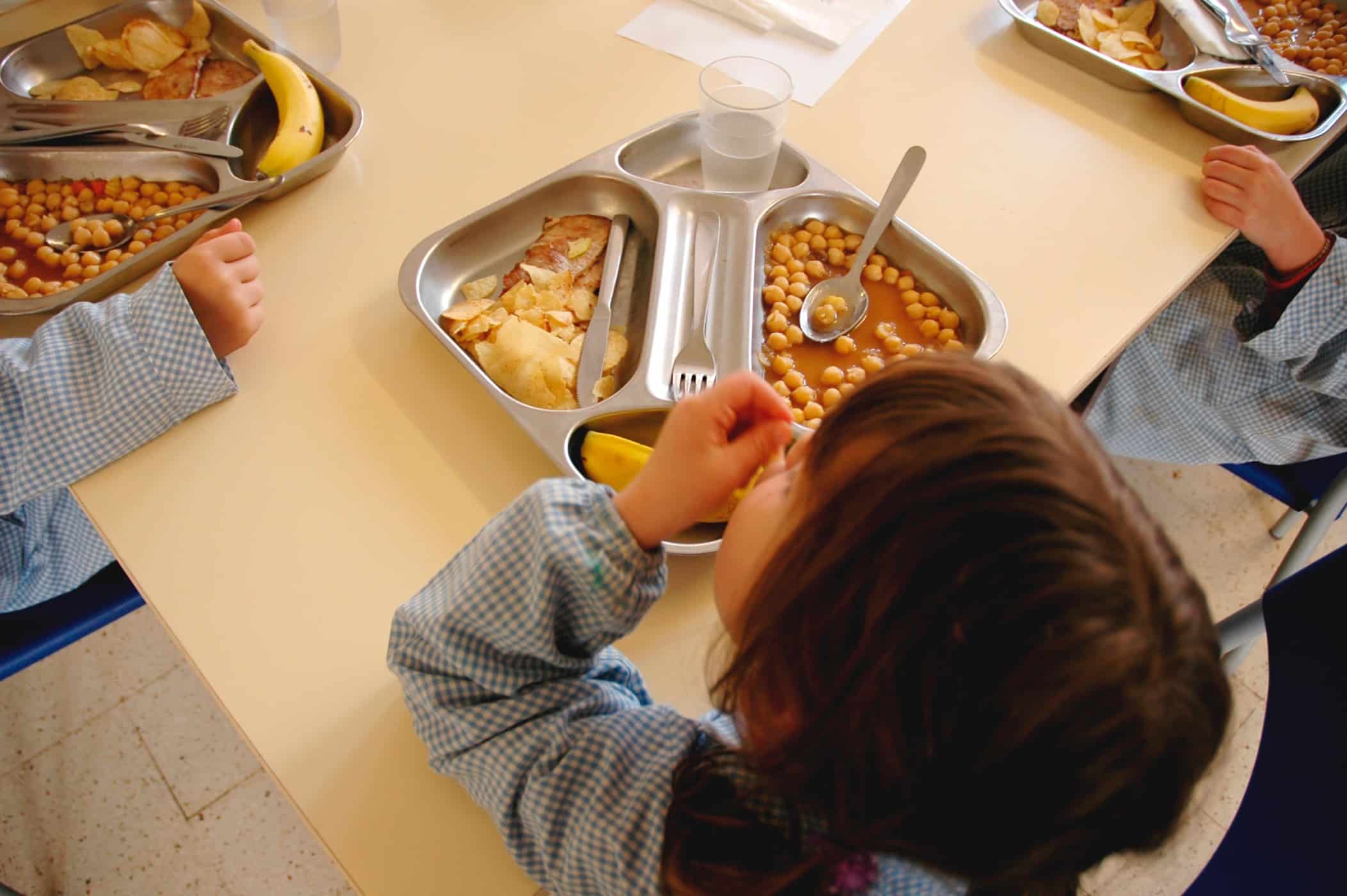 Un niño con una camisa azul a cuadros está sentado a una mesa con una bandeja metálica llena de comida, entre la que hay garbanzos, un plátano y agua. Cerca de allí se ve a otros niños, que también están comiendo platos similares.