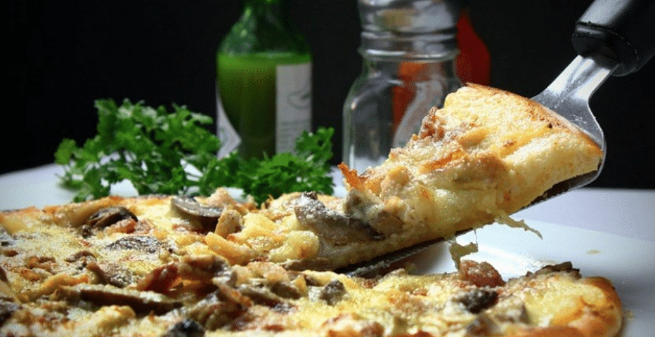 Una espátula saca de una pizza entera una porción de pizza con queso, champiñones y hierbas. Al fondo se ven perejil fresco, una botella verde y un salero.