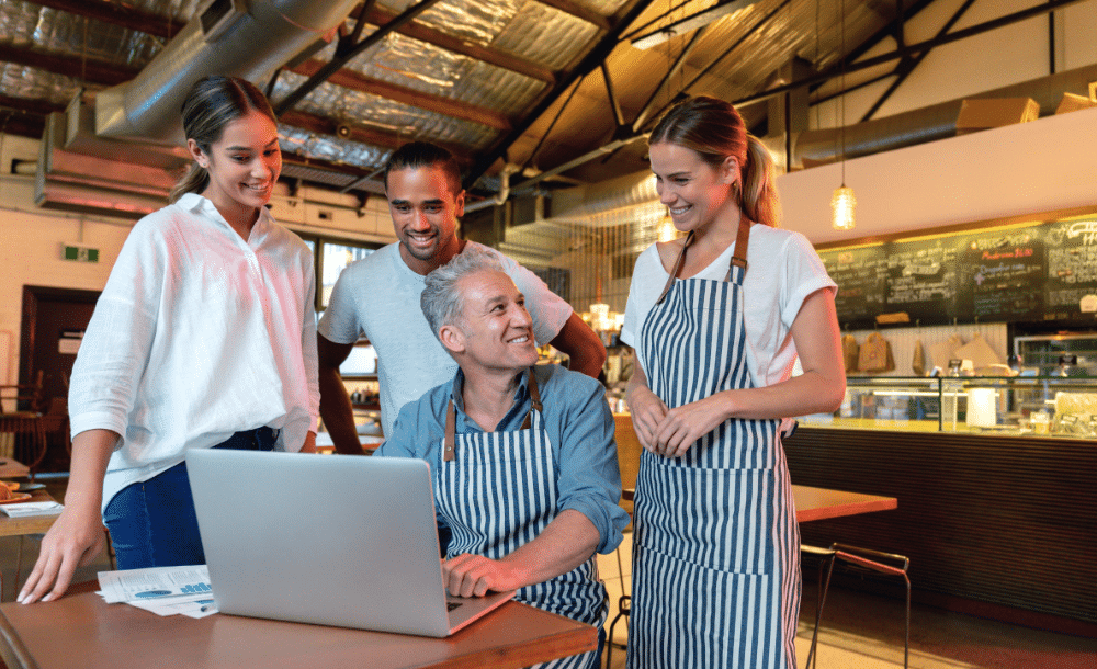 Cuatro empleados de un restaurante —tres de pie y uno sentado en una mesa con un ordenador portátil— sonríen y charlan en el ambiente informal y moderno de una cafetería. Dos de ellos llevan delantales a rayas.