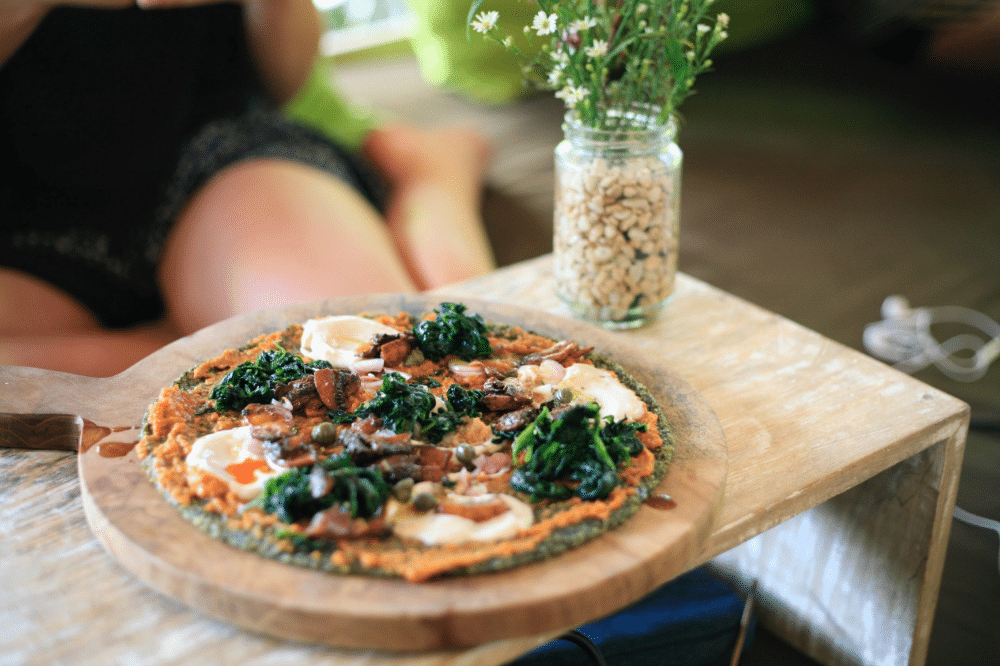 Una pizza recién hecha, con espinacas, champiñones y queso, descansa sobre una tabla de madera junto a un jarrón con flores y un tarro de piñones, con una persona sentada al fondo.