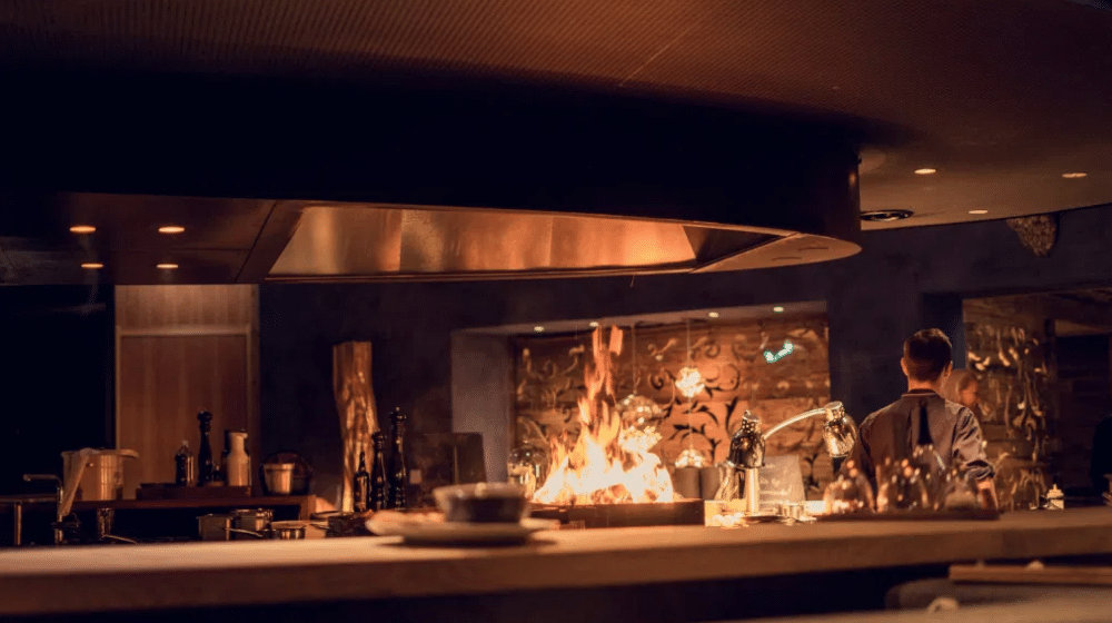 Un chef se encuentra en la cocina de un restaurante con poca luz, junto a una parrilla de llama abierta, con diversos utensilios y botellas sobre la encimera, lo que crea un ambiente cálido y acogedor.
