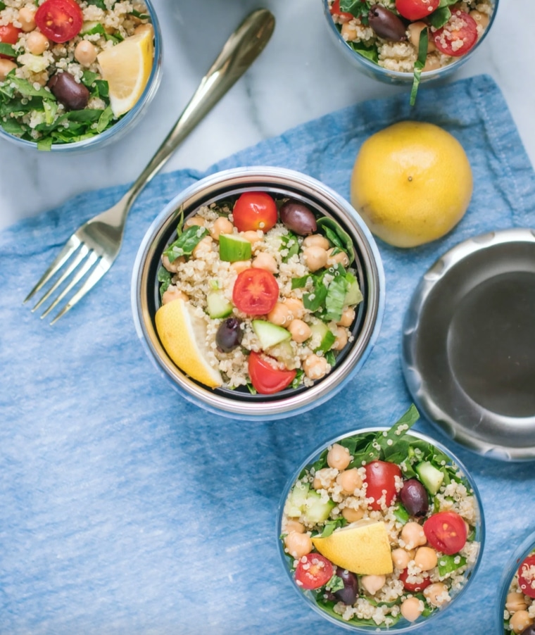 Ensalada de quinoa en tarros con tomates cherry, garbanzos, pepinos, aceitunas, espinacas y gajos de limón. Cerca, sobre un mantel azul, hay un tenedor, una tapa de metal y un limón.
