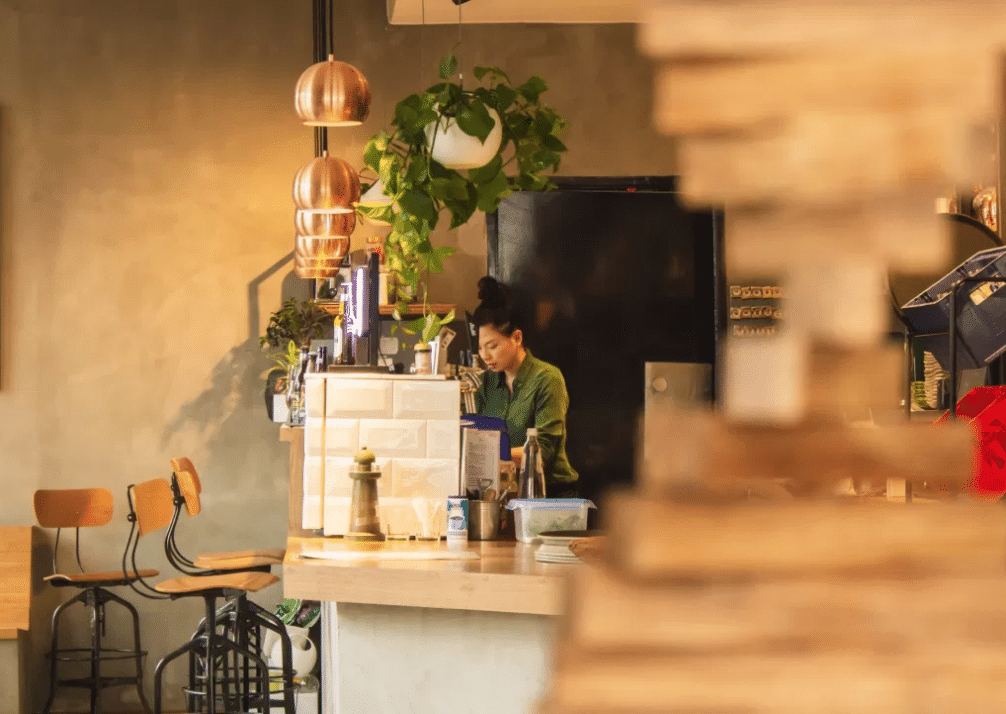 Una persona está de pie detrás de la barra de una acogedora cafetería con sillas de madera, lámparas colgantes y una planta en el techo. La escena está iluminada con una luz cálida y resulta muy acogedora.