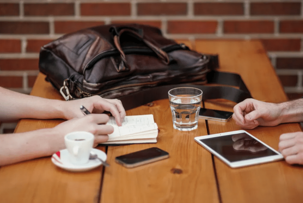 Dos personas están sentadas ante una mesa de madera con un cuaderno, un bolígrafo, un smartphone, una tableta, una taza de café, un vaso de agua y un bolso de piel. Solo se les ven las manos.