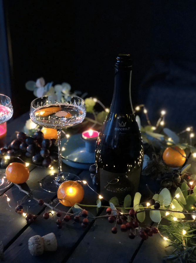 Una mesa festiva con una botella de vino espumoso, dos copas, uvas, mandarinas, velas, corchos y guirnaldas de luces que crean un ambiente acogedor.