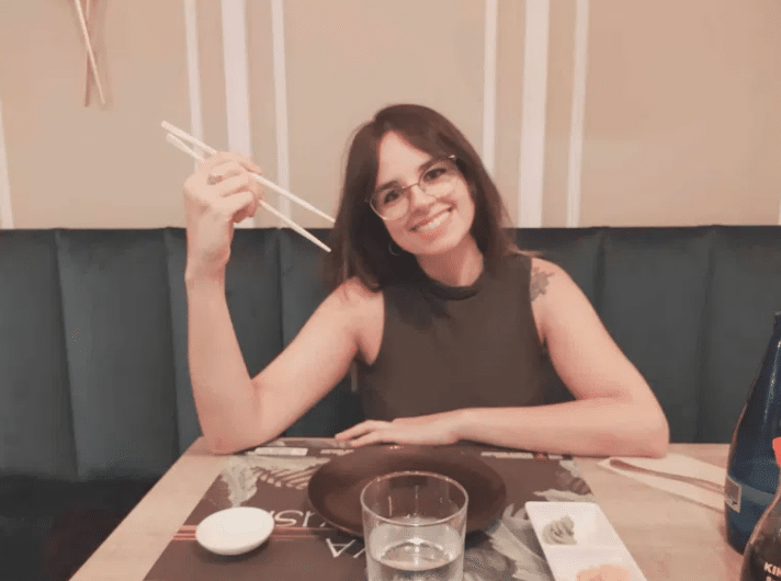 Una mujer con gafas sonríe sentada a la mesa de un restaurante, con los palillos en una mano. Sobre la mesa hay un vaso de agua, un plato y unos cuencos pequeños con wasabi y jengibre. Parece relajada y feliz.