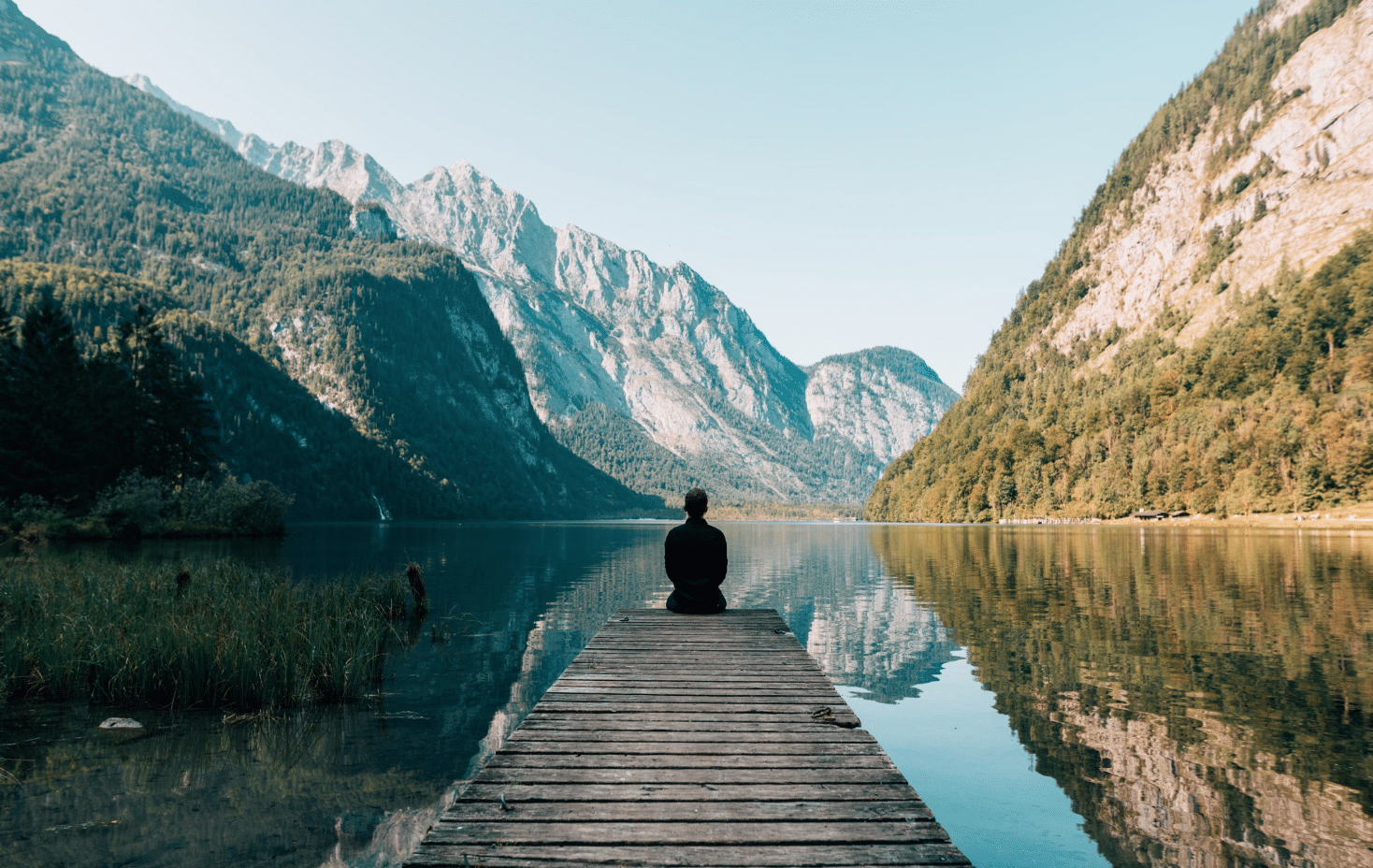 Una persona está sentada al final de un embarcadero de madera, con vistas a un lago en calma rodeado de altas montañas verdes, bajo un cielo azul y despejado.