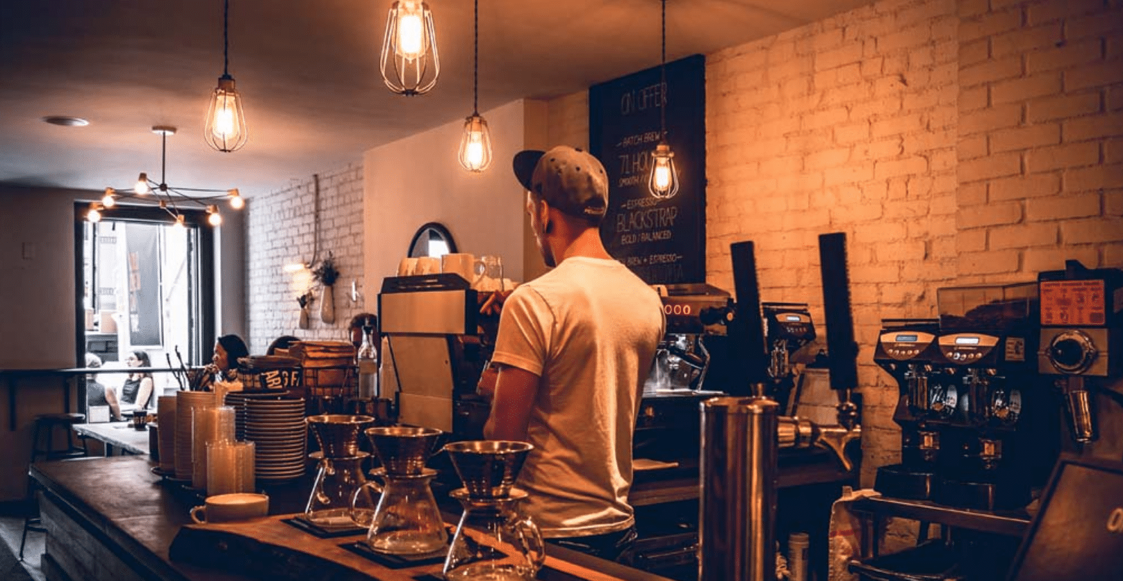 Un barista está detrás de la barra de una cafetería acogedora y con una iluminación cálida, preparando bebidas. Se ven mesas, el equipo de café y una pizarra con el menú. Un cliente está sentado en una mesa junto a la ventana.