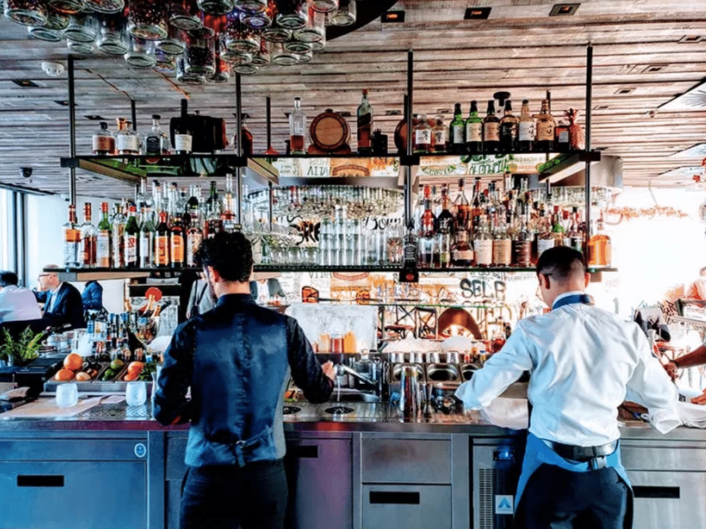 Dos camareros trabajan detrás de la barra de un bar muy concurrido, repleta de botellas y vasos. En las estanterías se ven diversas botellas de licor. El ambiente es animado y, al fondo, se divisan otros clientes.