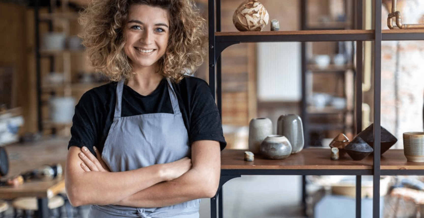 Una persona sonriente, con el pelo rizado y un delantal gris, está de pie con los brazos cruzados en un taller de alfarería, con estanterías en las que se exponen piezas de cerámica al fondo.