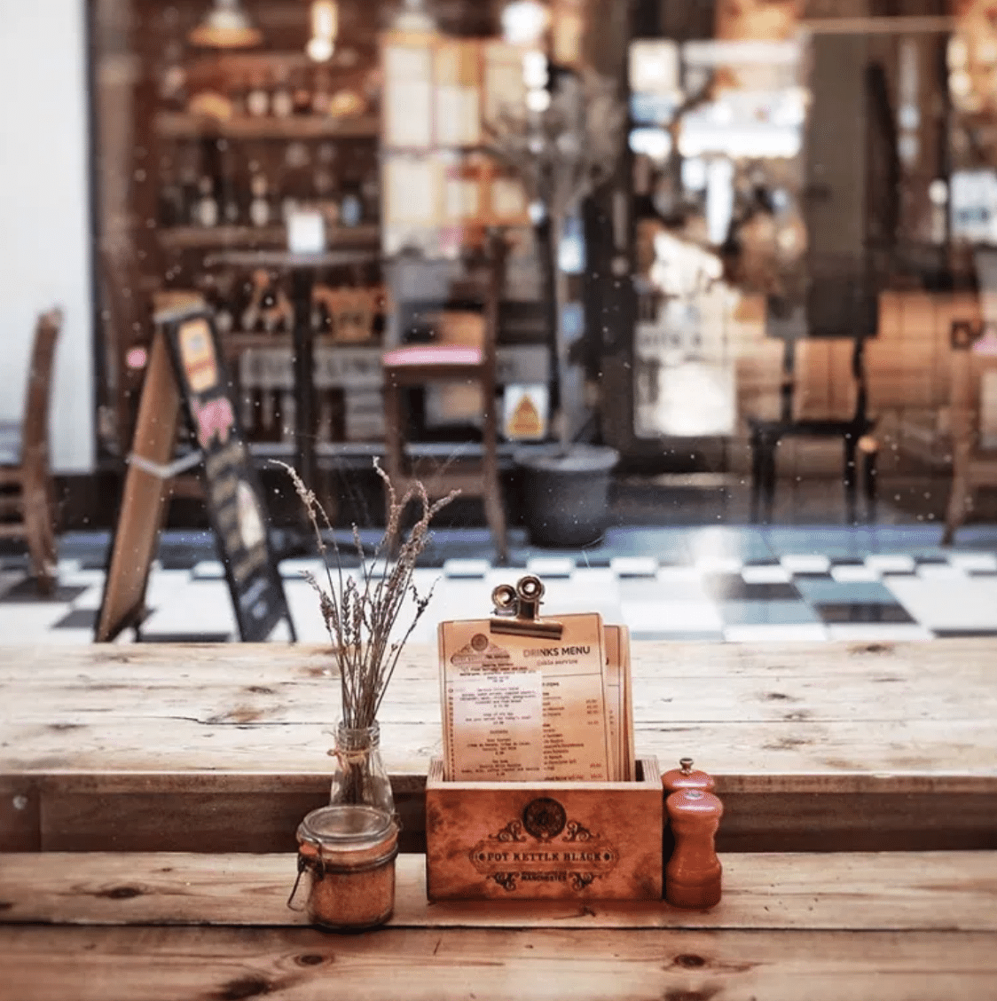 Sobre una mesa de madera hay un menú, flores secas en un jarrón, un tarro de cristal pequeño y un molinillo de pimienta roja; al fondo se ve el interior de una cafetería a través de una ventana.