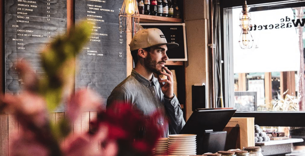 Un hombre con una chaqueta gris y una gorra está de pie detrás del mostrador de una cafetería, con aire pensativo. Al fondo se ven estanterías con botellas y un cartel con el menú. En primer plano hay unas flores borrosas.