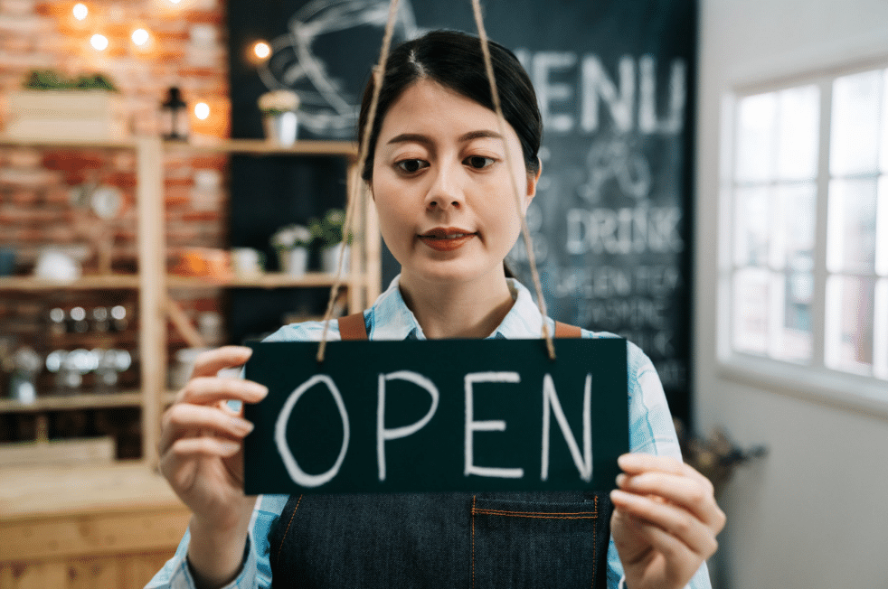 Una mujer con delantal cuelga un cartel que dice «ABIERTO» en una acogedora cafetería, con una pizarra con el menú y unas cálidas luces visibles al fondo.