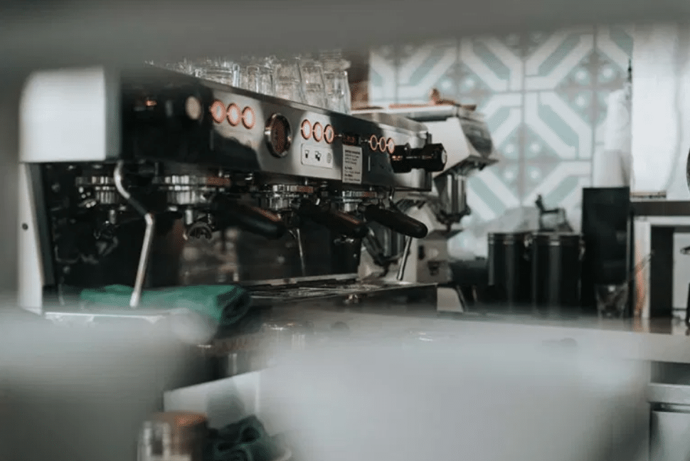 Primer plano de una máquina de café espresso profesional con dos portafiltros y vasos encima, situada en una cafetería con azulejos estampados al fondo.
