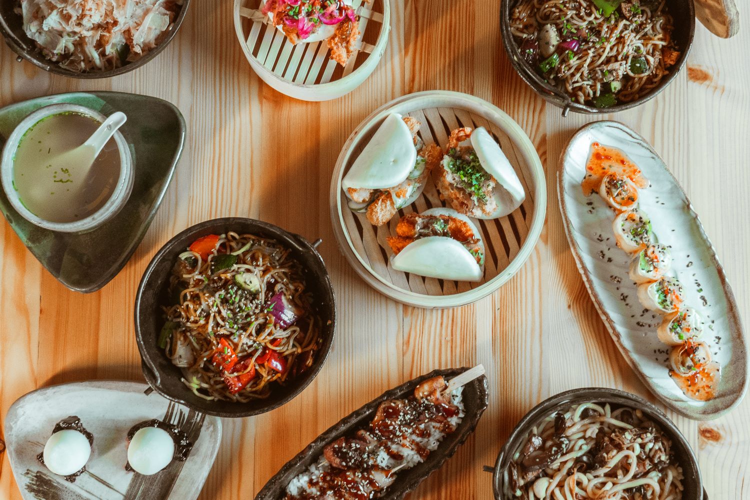 Sobre una mesa se exponen diversos platos asiáticos, entre los que se incluyen fideos, bollos bao, sopa, empanadillas, rollitos de sushi y postres, dispuestos ordenadamente en platos de madera y cerámica.