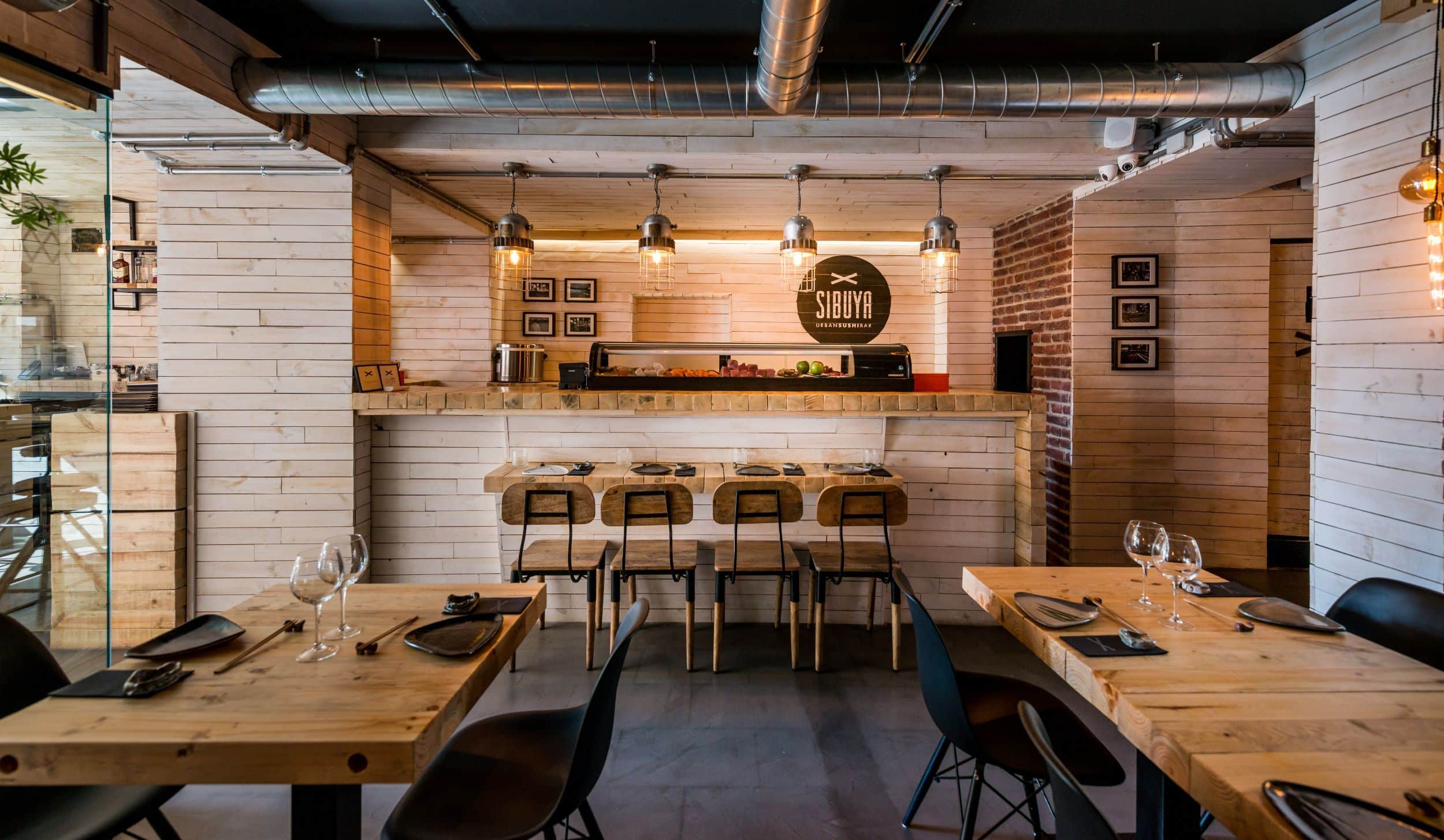 Interior de un restaurante de sushi moderno con mesas de madera, sillas negras, una barra con taburetes, lámparas colgantes y conductos a la vista. Los cubiertos y las copas de vino están dispuestos de forma ordenada sobre las mesas y la barra.